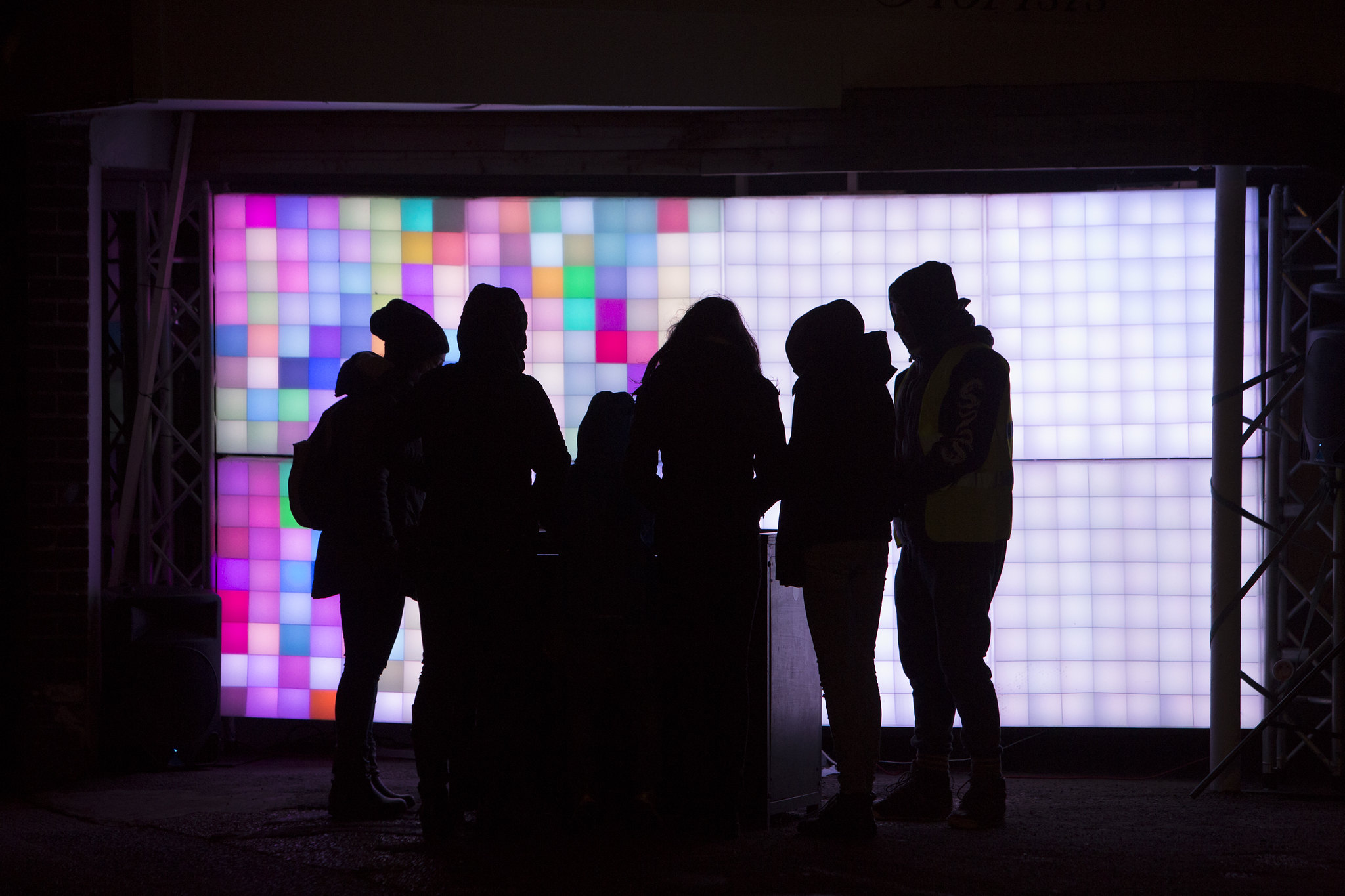 A group of children and adults stand around a table, with a colourful wall of LED lights behind them