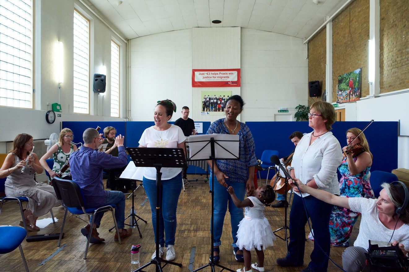 Two women are standing in front of musicians in a community hall setting, singing at music stands, with a woman kneeling and recording the music, and a child trying to get one of the singer's attention