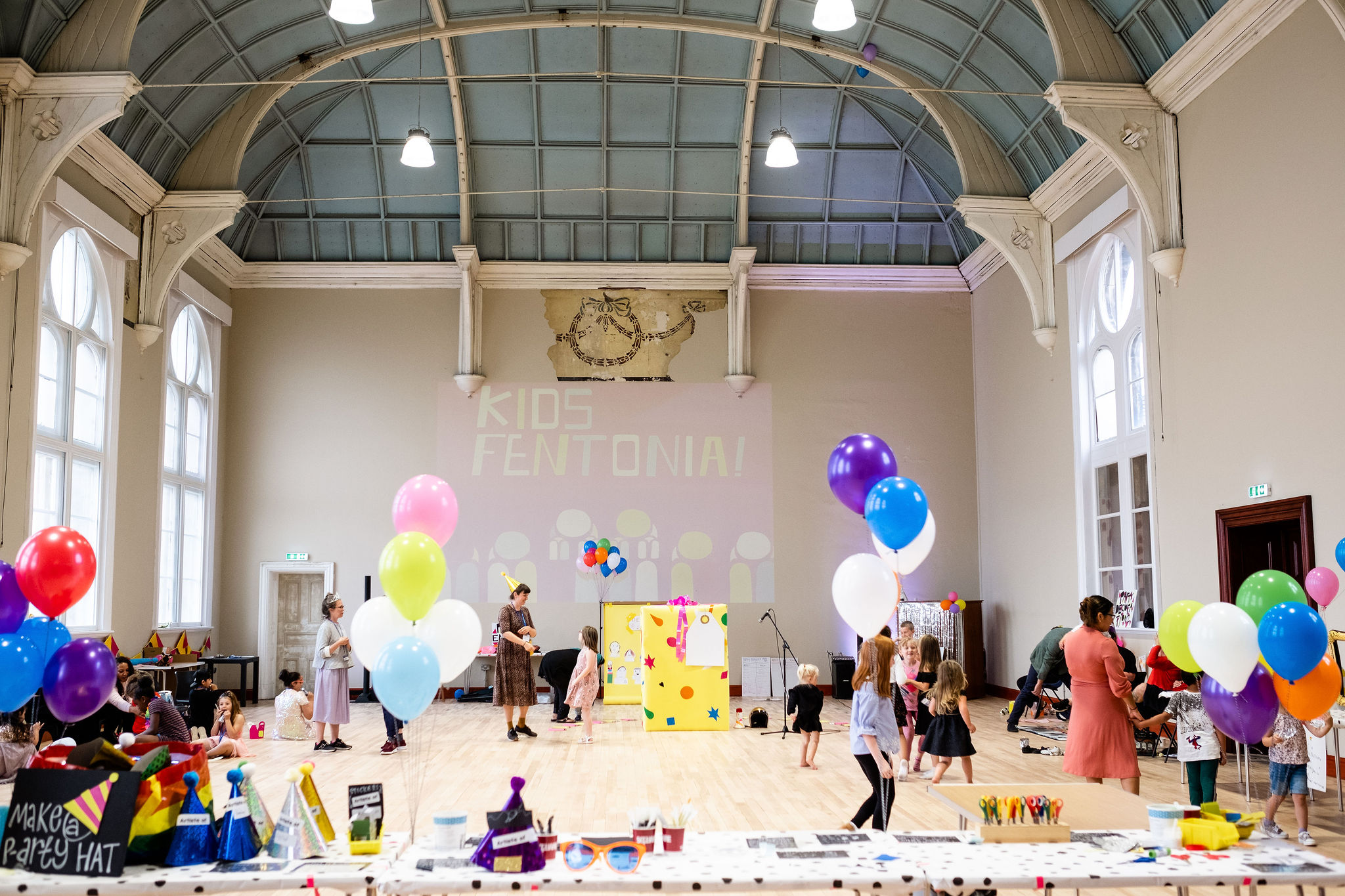 Children play in a large room with vaulted ceiling, wearing colourful party hats and surrounded by balloons. 