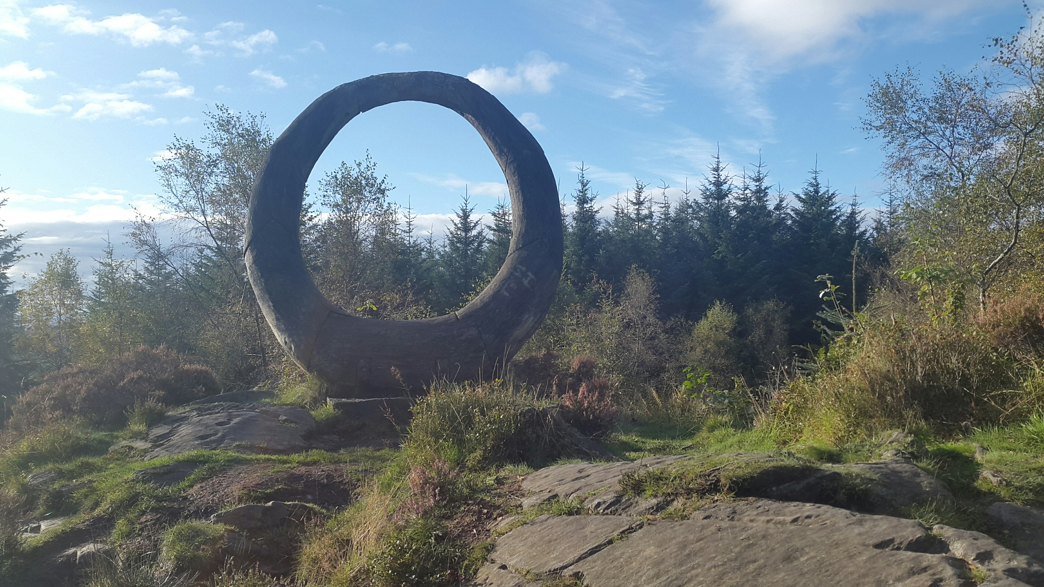 A photograph of a sculpture in an outdoor location. A large, round form is shown in front of trees. It appears to be made from stone.