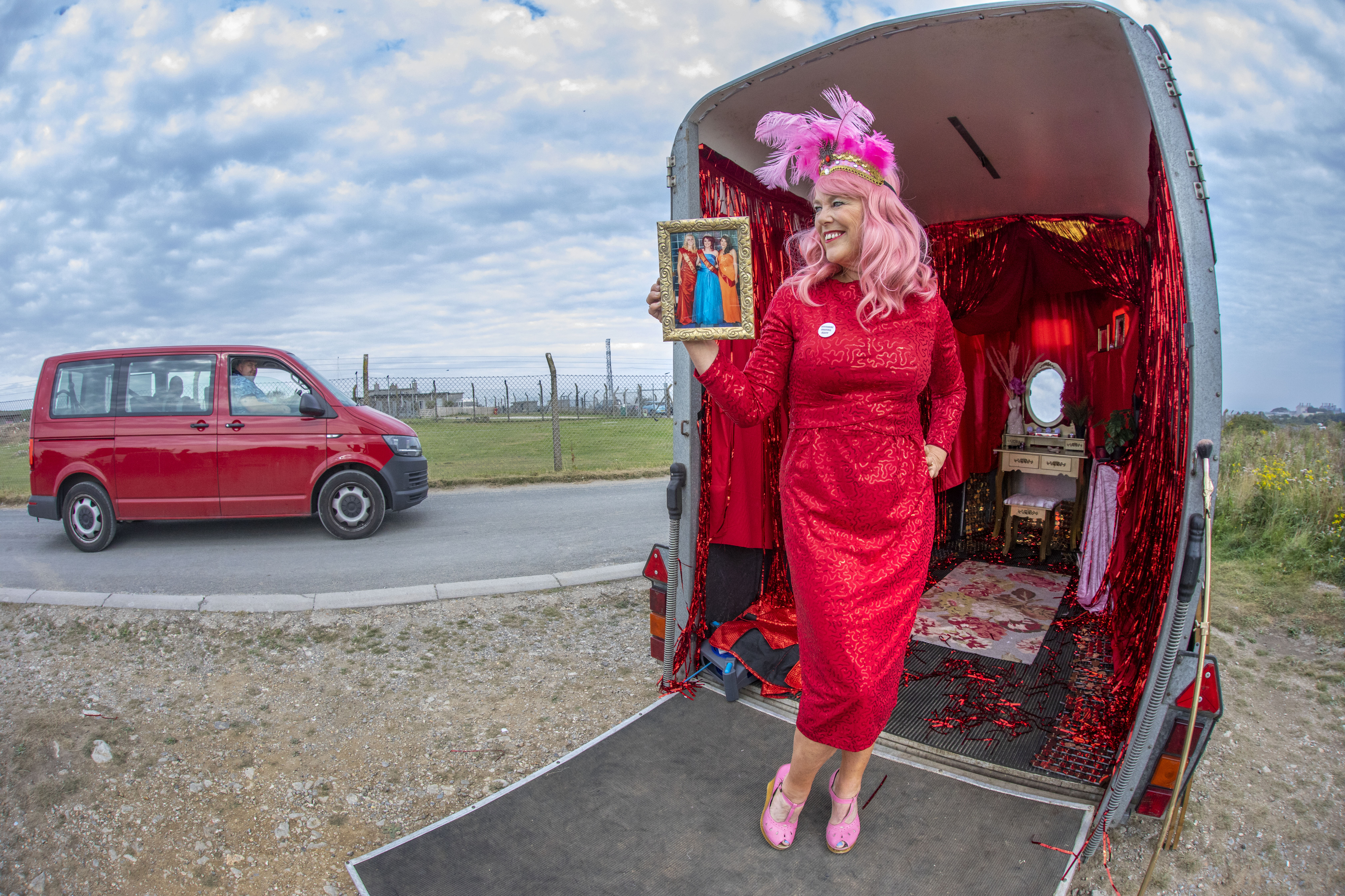 In an outdoor setting the side of a road, a woman, dressed head to toe in red, wearing a pink wig and makeup, stands next to a horse box. The inside of the horsebox is ellaborately decorated in shades of red and pink.