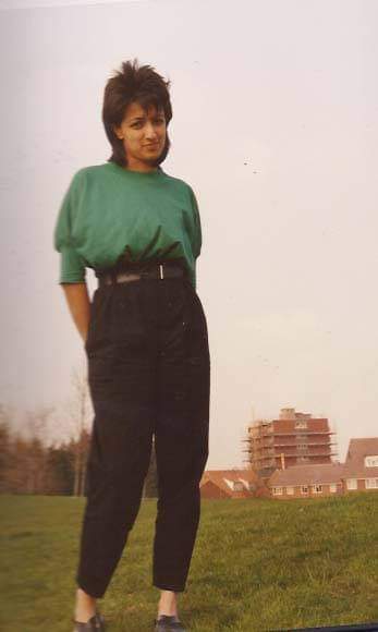 Photograph of a young woman with short dark hair, wearing a green top and high-waisted trousers. Her hands are in her pockets and she looks straight at the camera.