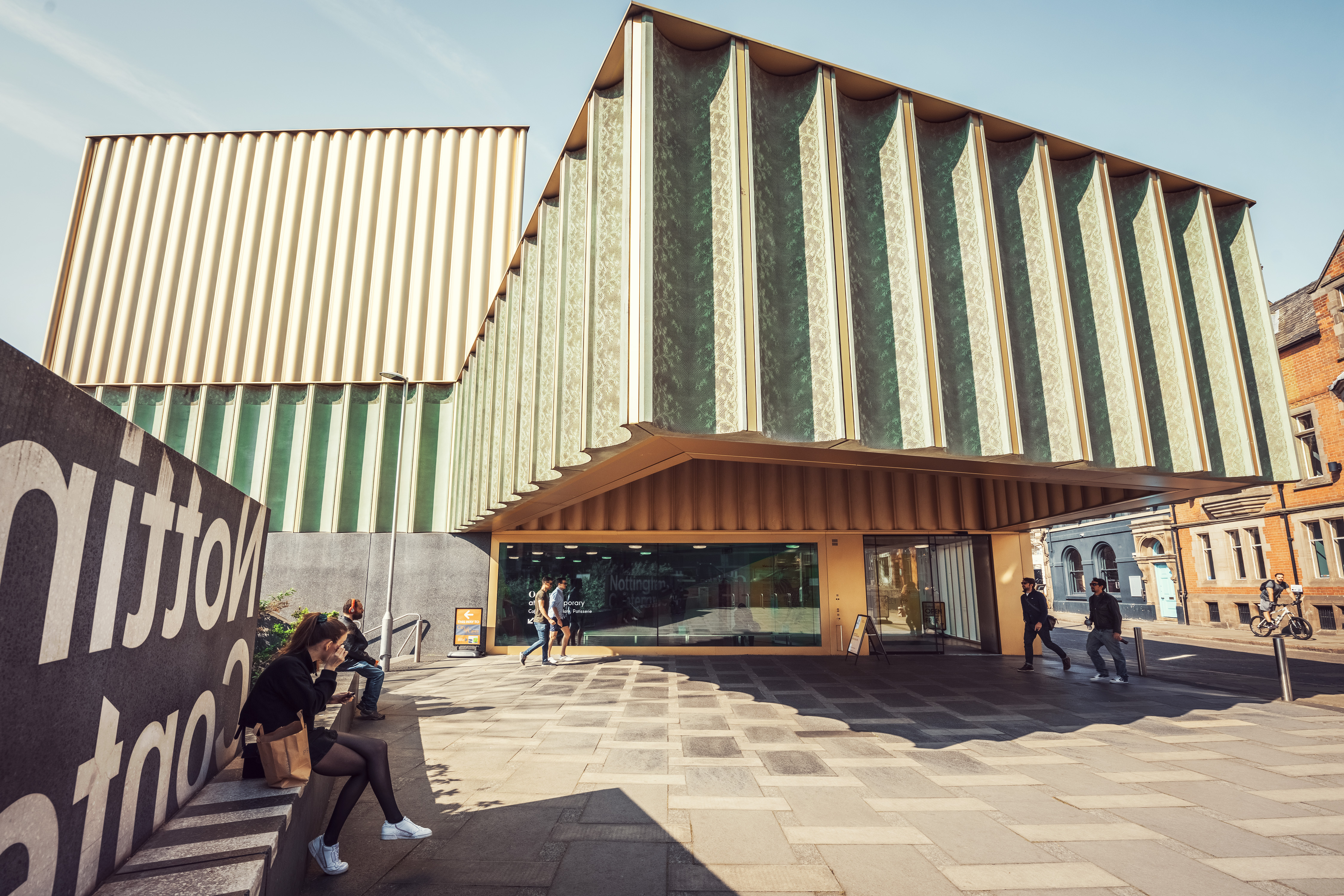 The entrance to Nottingham Contemporary's iconic angular green and gold building, with visitors sitting and walking outside.