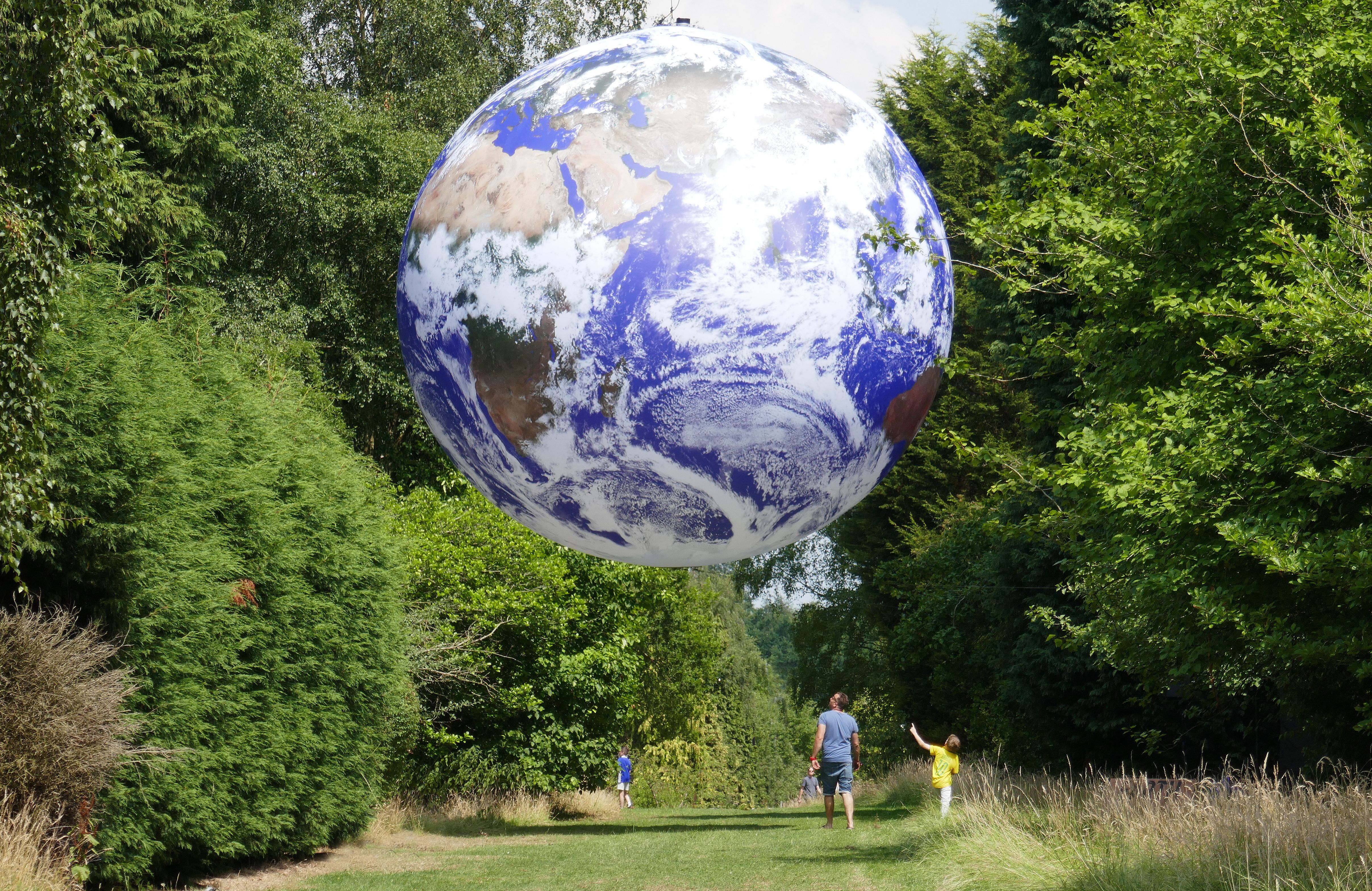 In an outdoor setting, an adult and child look up above them to see an enormous globe that is positioned in the sky, reaching the height of the tops of the trees.