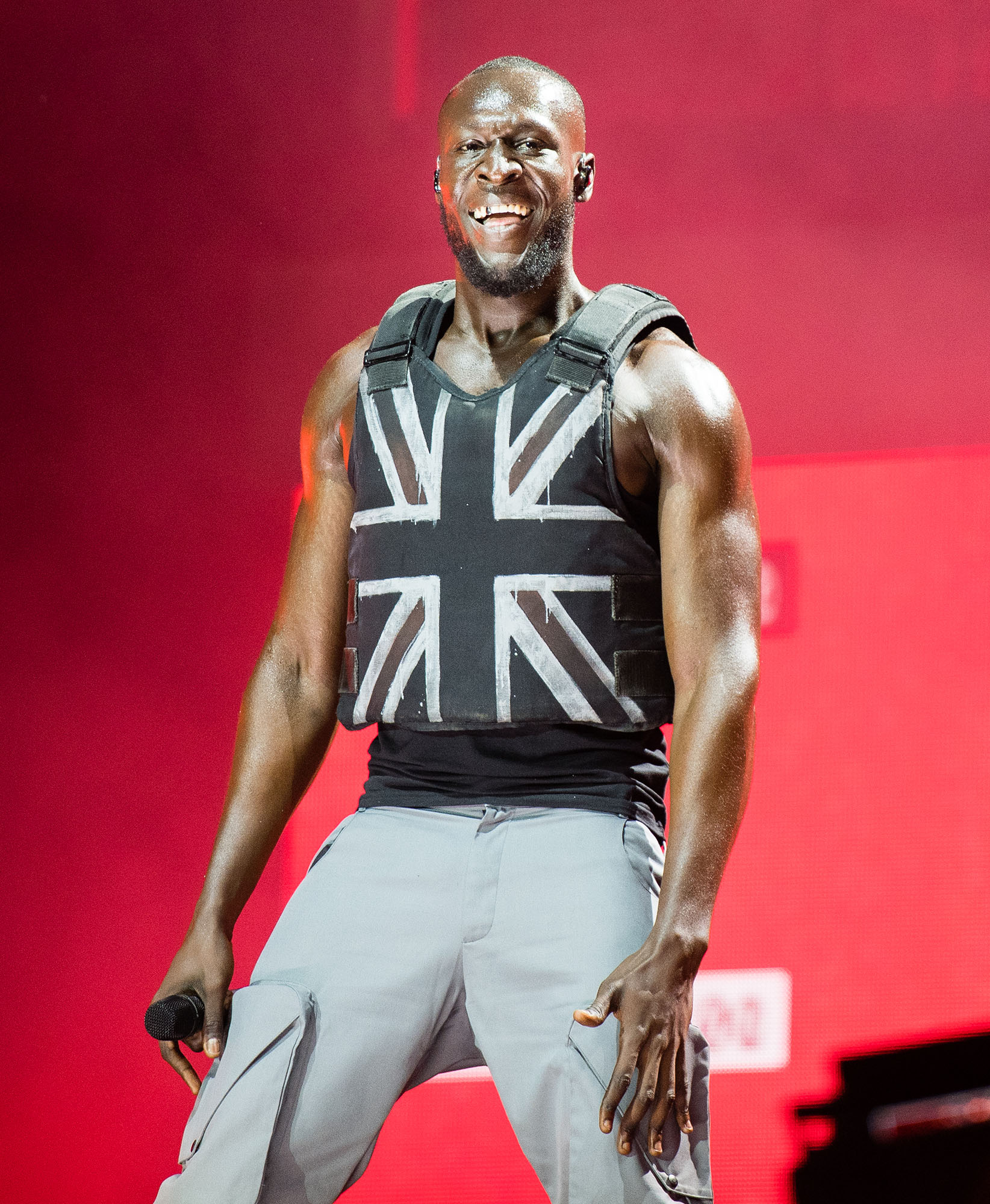 Stormzy performs onstage at Glastonbury festival. He is holding a microphone and wears a stab proof vest with the union jack painted on the front.