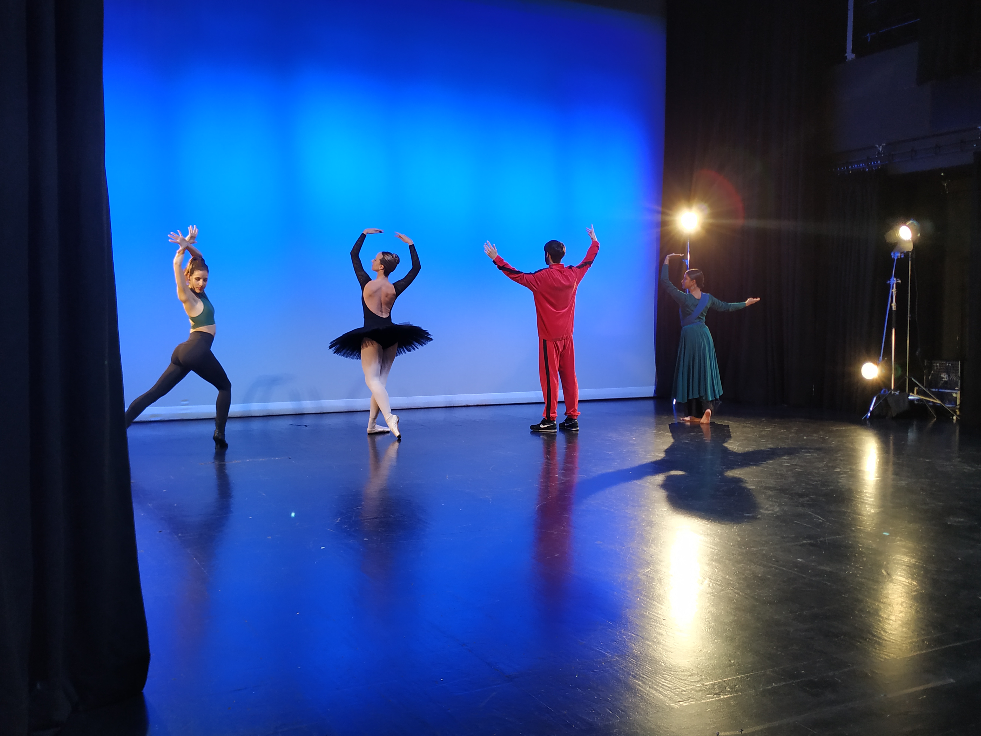 Four dancers rehearse on an empty black stage, with bright blue lights. They wear grey, black, red and blue costumes.