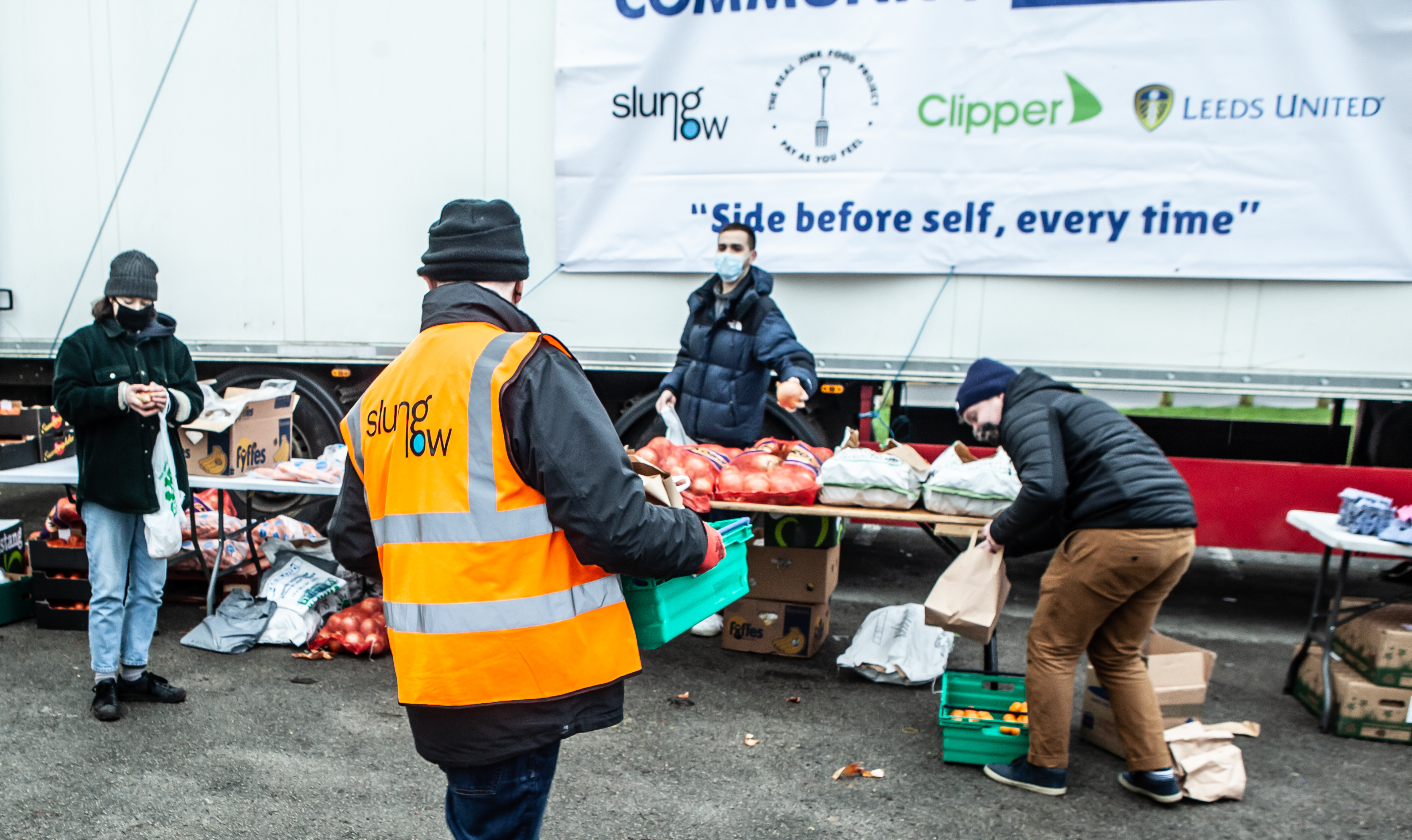 A person carries a crate of food packages to a table at an outdoor foodbank.