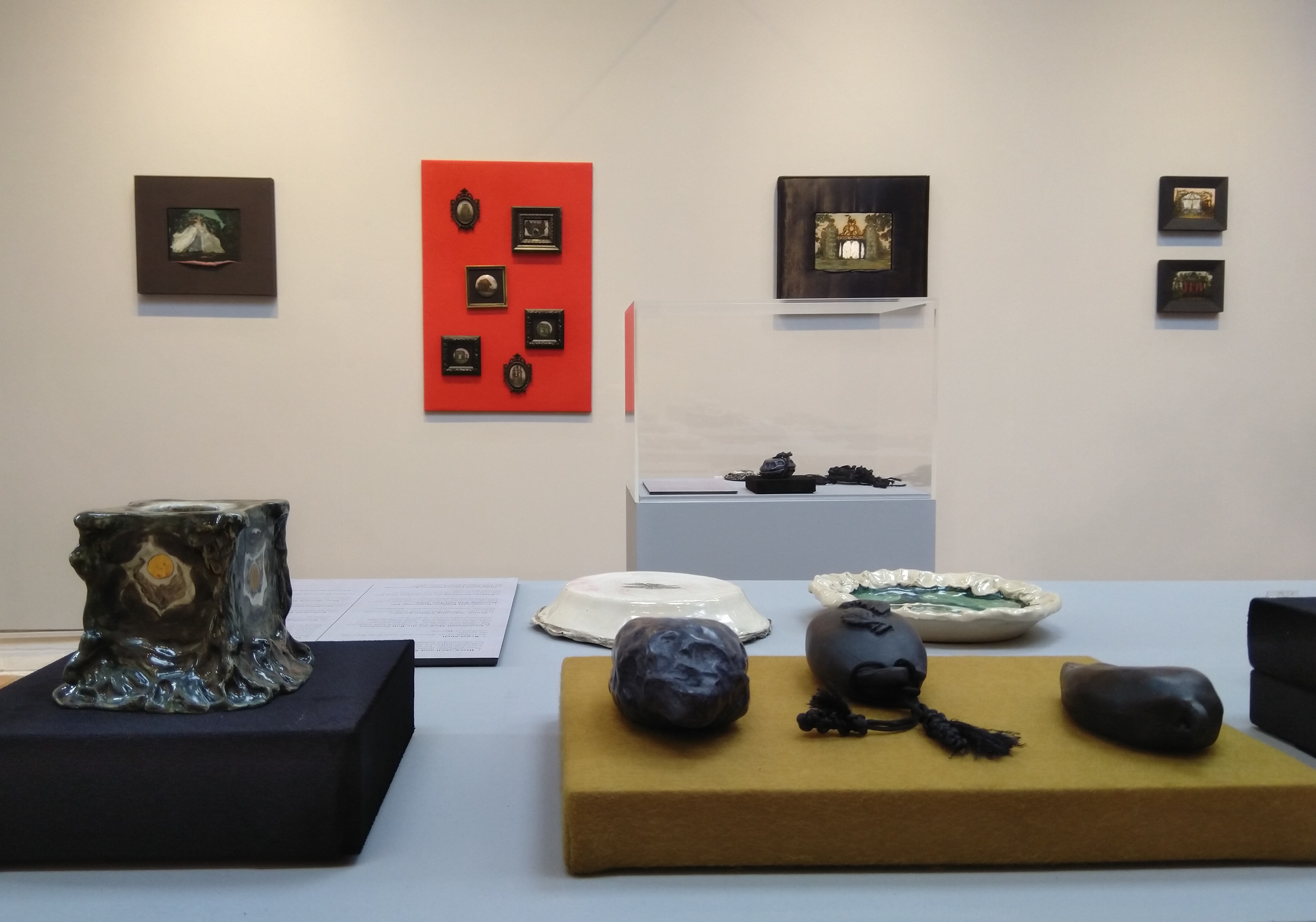Inside the exhibition, showing a table with six ceramic objects, a glass cabinet and five paintings on the wall.