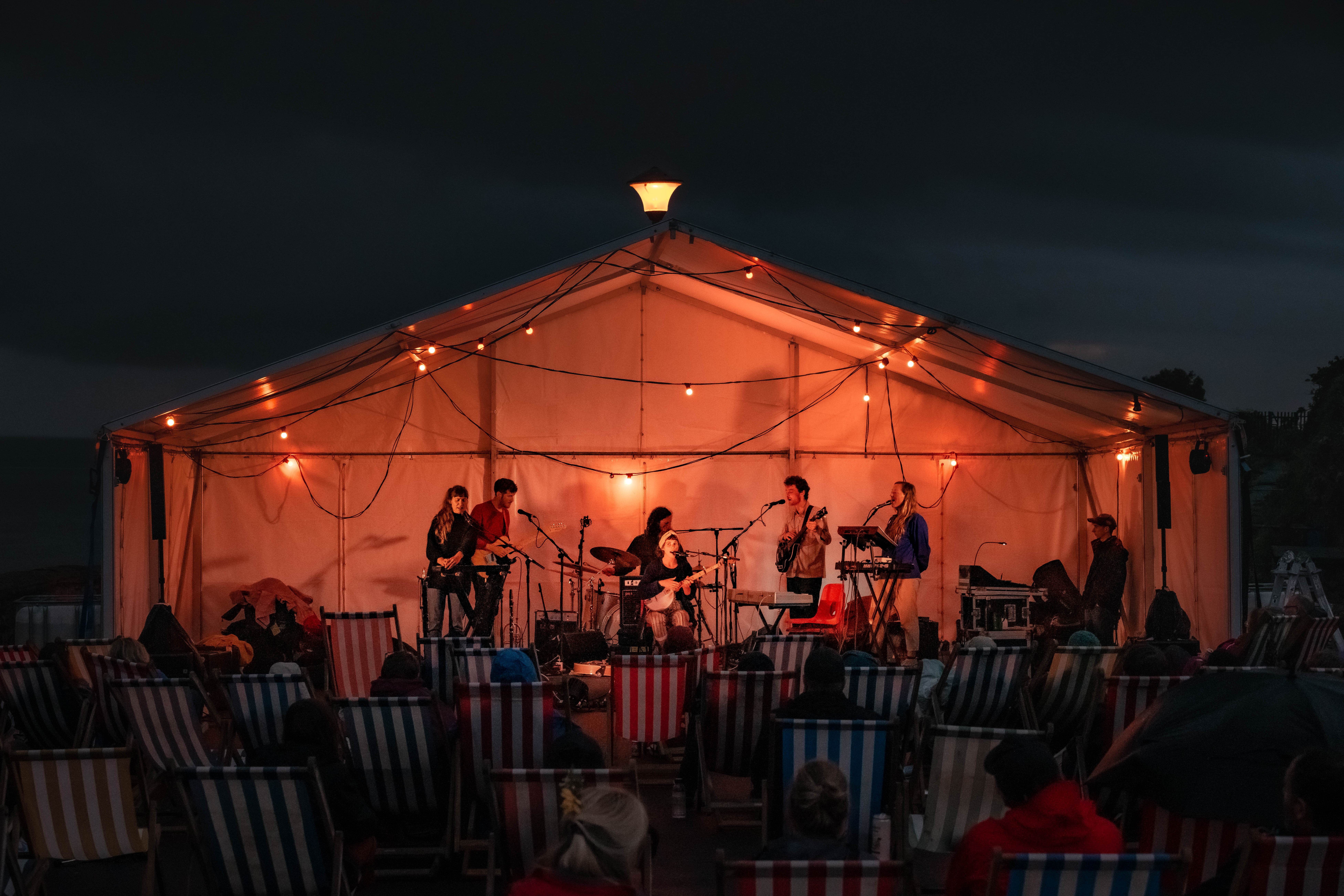 Musicians are performing under a tent at night, lit up with orange lights. They are performing in front of an audience sat on deckchairs 