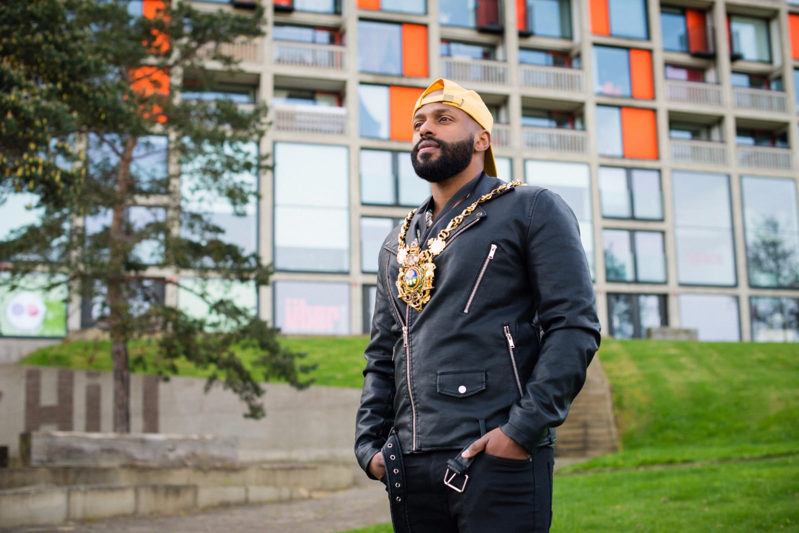 A person in a yellow baseball cap and gold medallion chains stands in front of block of flats 