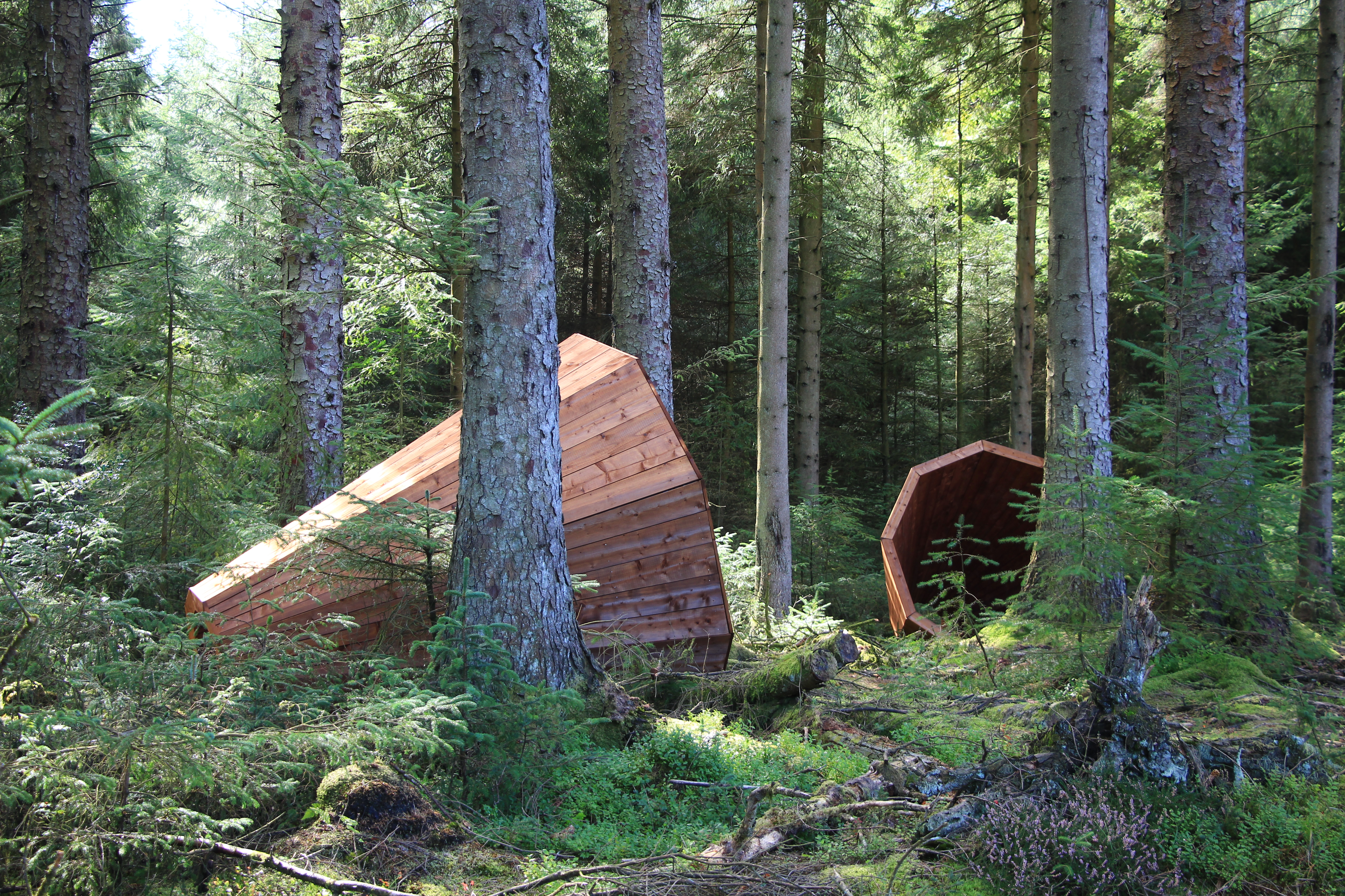 A photograph of two wooden sculptures within a forest. The sculptures are in a cone shape.