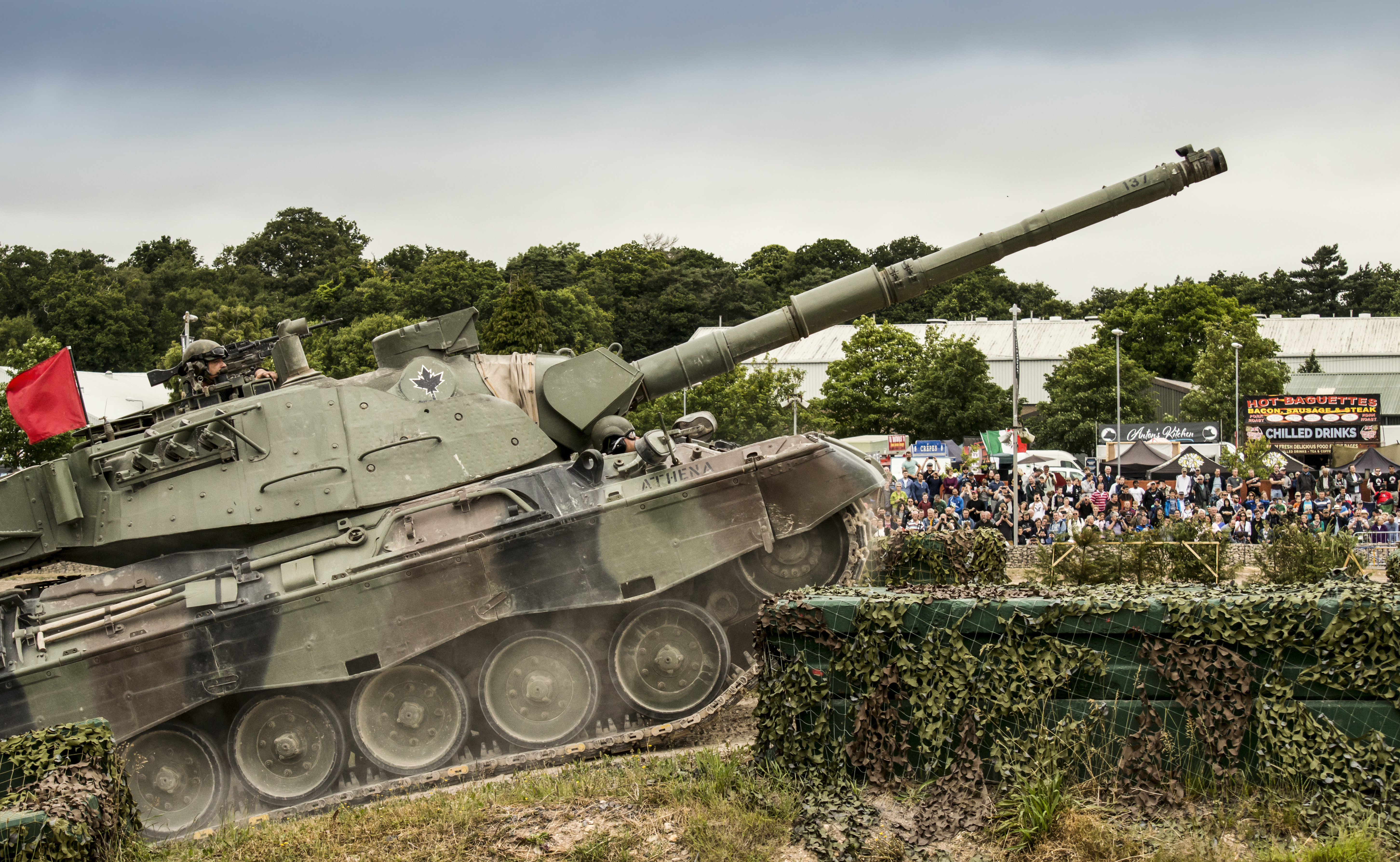 In an outdoor setting, an army tank is driving across the view. People are watching and there are festival stalls in the background.