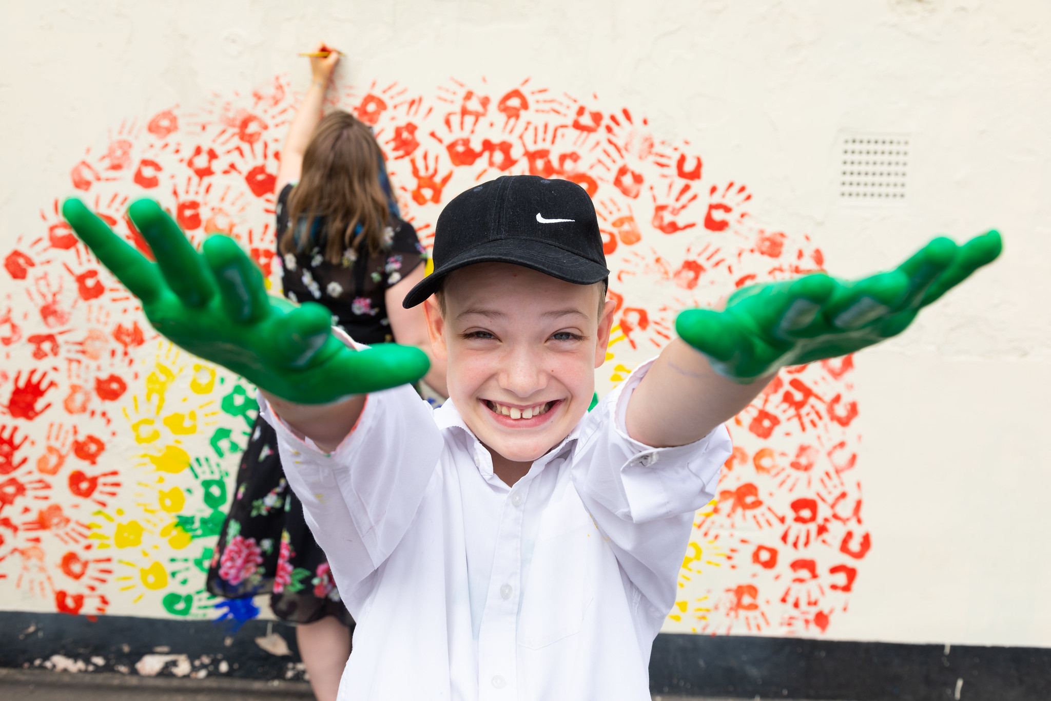 A young person with green paint on their hands stands in front of a wall covered in a multicoloured hand prints