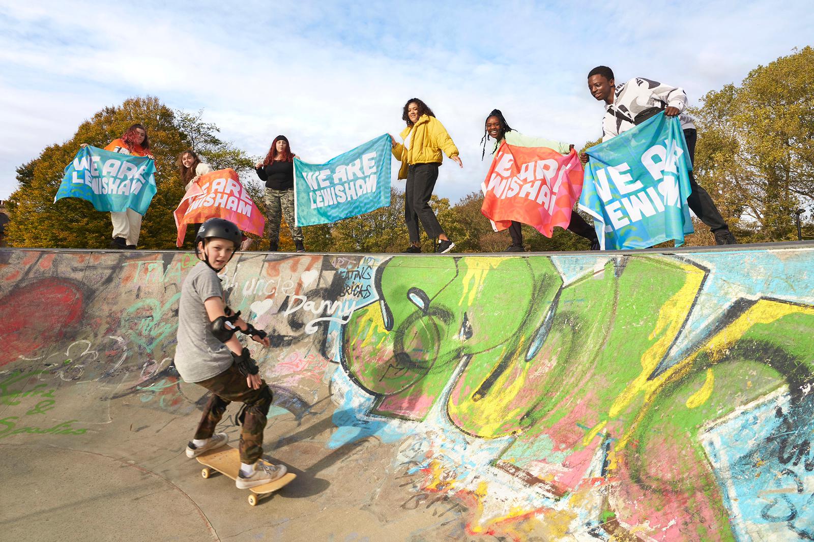 A photograph of a group of people at a skate ramp. They hold signs that read: We Are Lewisham. In the foreground, a person wearing a helmet is shown on a skateboard.