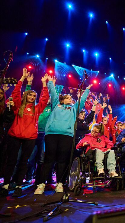 A group of young people stand on a stage in colourful sweatshirts doing a live performance of musical instruments. 