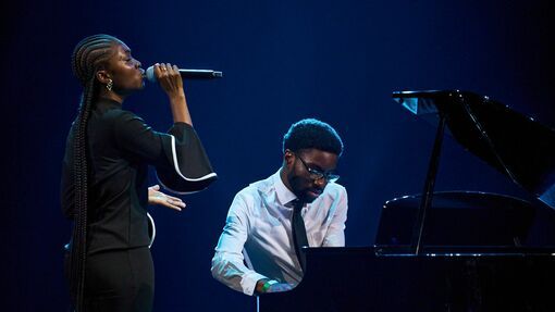 A young black woman stands on stage, with a microphone singing. She is accompanied by a young man sat on a piano next to her, wearing a white shirt. 