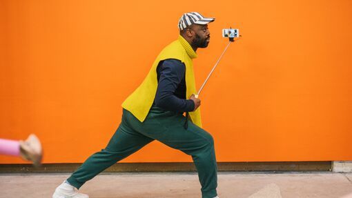 Artist Harold Offeh photographed in a lunge pose while holding a phone camera on a selfie stick in front of an orange wall 